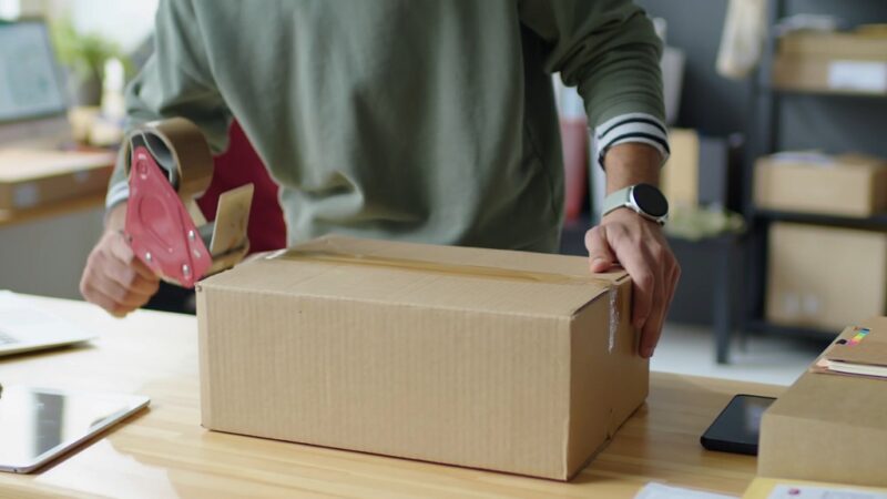 Close-up of a person taping a cardboard box for shipping, highlighting unoptimized packaging contributing to business overhead