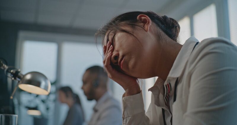 Office worker holding her head in frustration, with coworkers in the background, depicting the cost of poor scheduling on business overhead