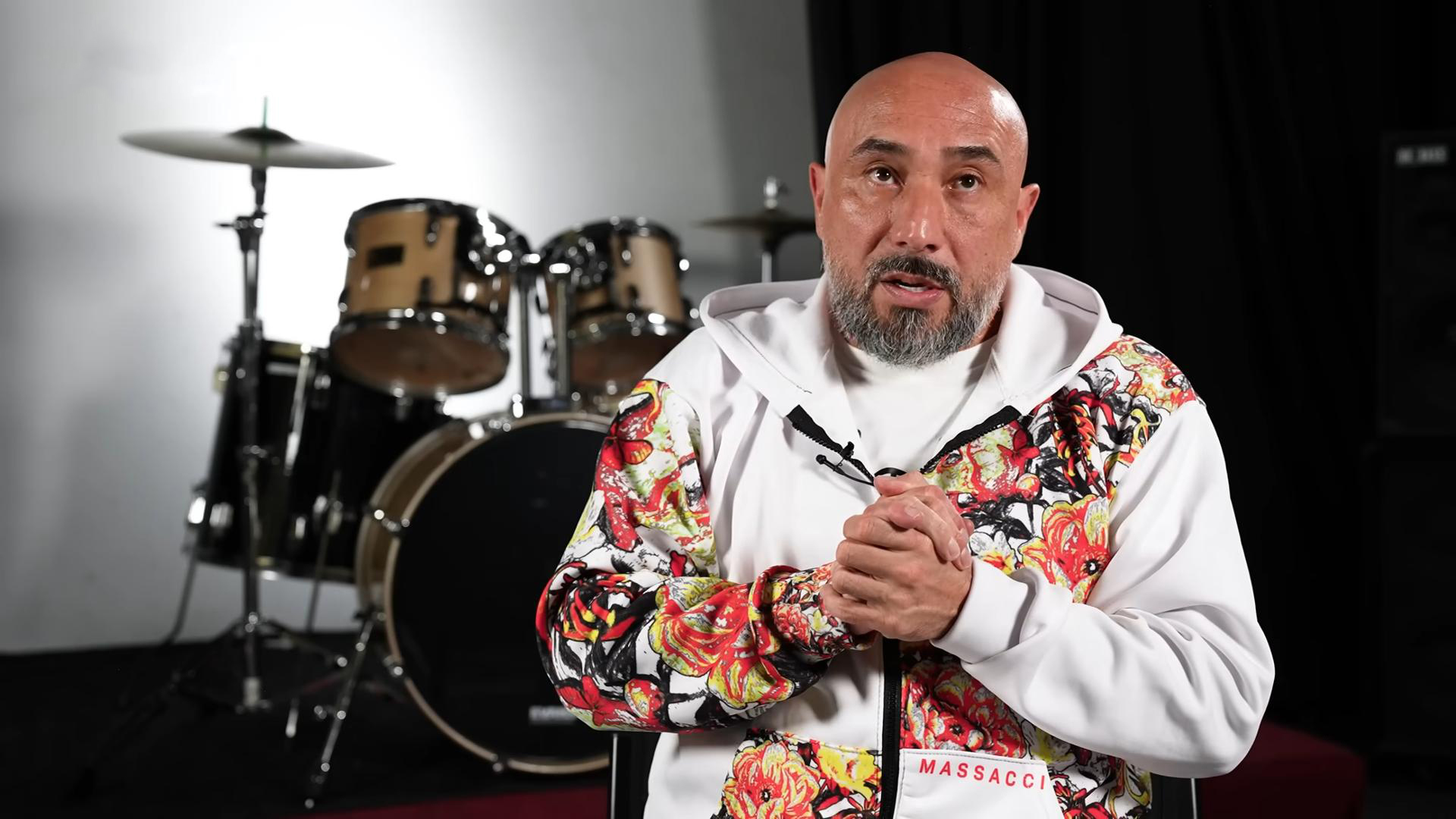 Man in a white floral hoodie speaking while seated in front of a drum set