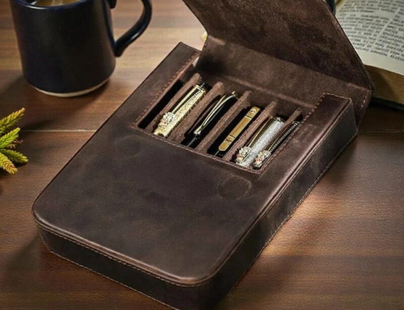 Open dark brown leather pen case with multiple pens neatly arranged inside, placed on a wooden desk next to a coffee mug and an open book