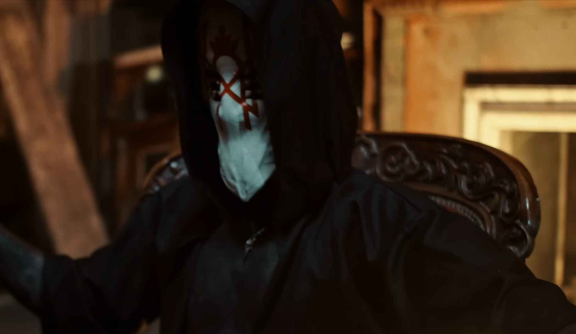 Masked and hooded performer seated in a dimly lit room