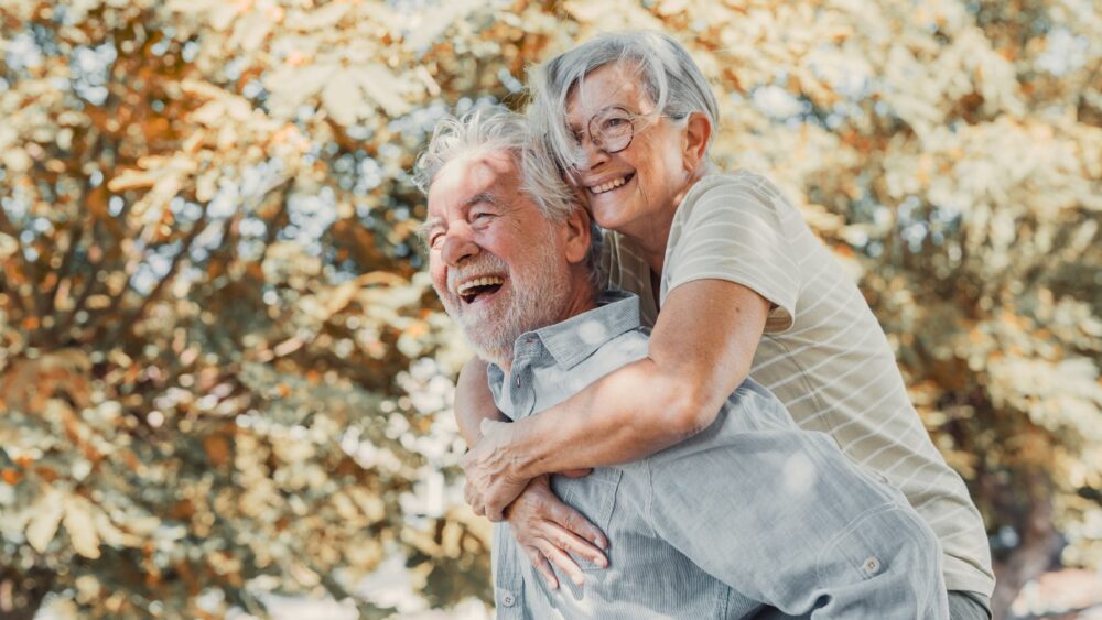 Smiling older couple outdoors, with a woman hugging a man from behind in a sunlit park.