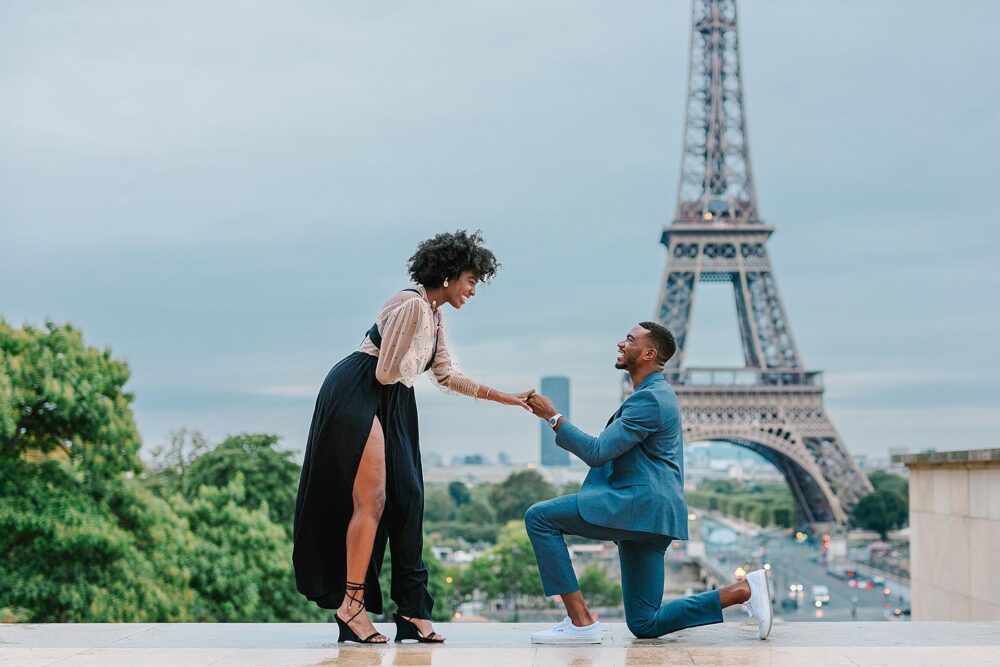 A man kneels to propose to his partner on a terrace with the Eiffel Tower in the background.