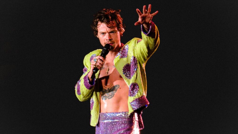 Performer on stage wearing a bright sequined jacket and high-waisted pants, illustrating bold gender-fluid fashion and expressive performance style.