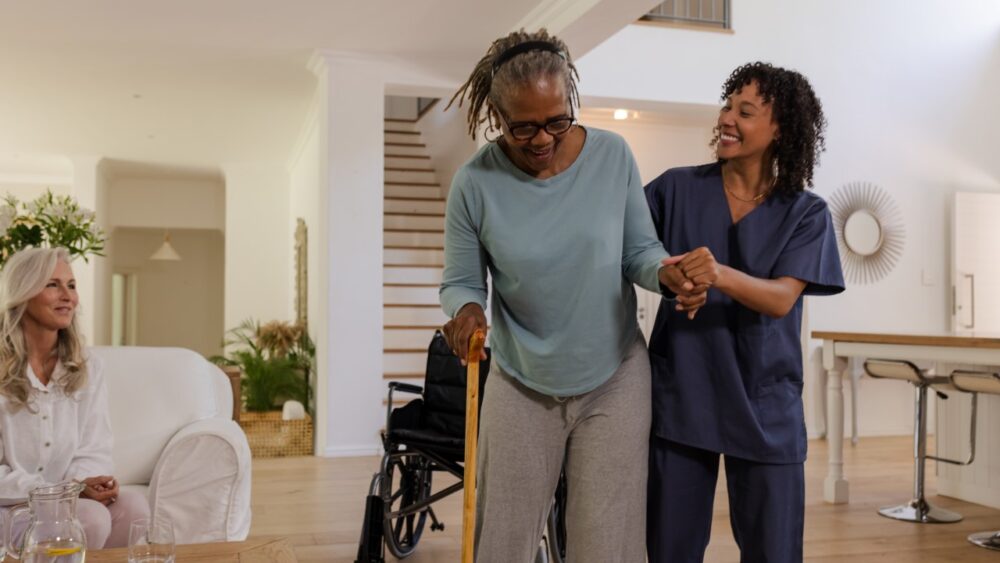 Professional caregiver helping an older woman walk with a cane in a bright home while another woman watches nearby.