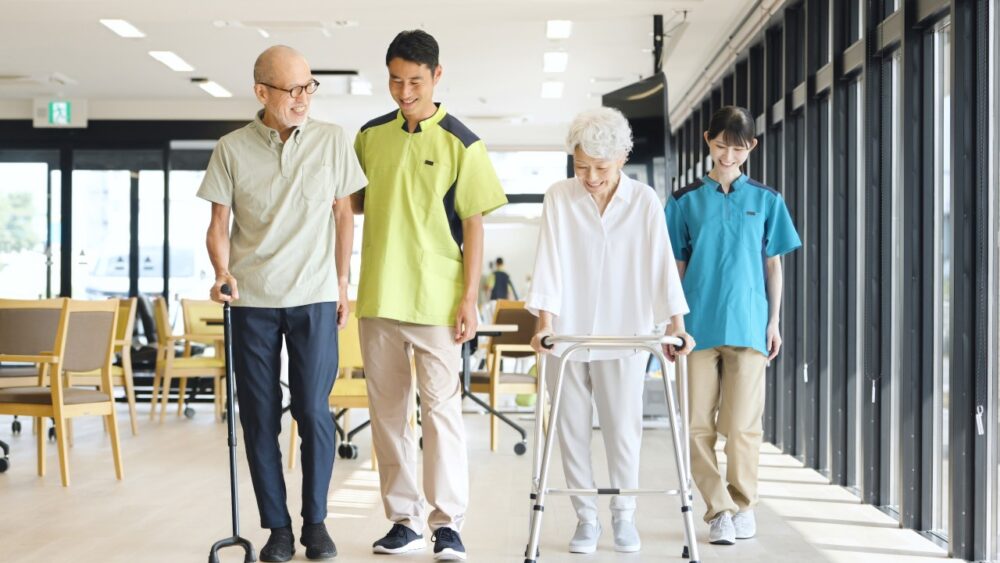 Two caregivers walking with an older man using a cane and an older woman using a walker in a care facility hallway.