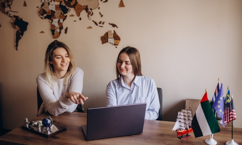 Two women are sitting at a desk with a laptop, smiling and engaged in discussion. A world map decorates the wall, and small flags and a model ship sit on the desk