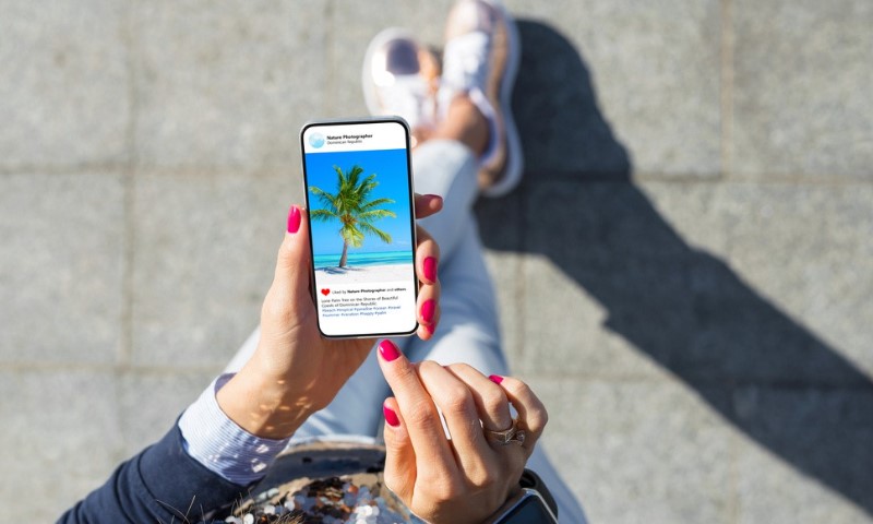 A person with pink nails and casual clothes is seated outdoors, browsing a beach photo on a smartphone