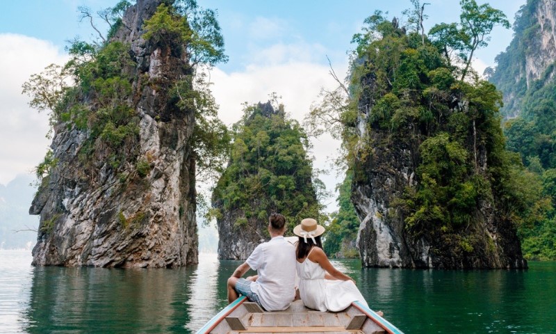 A couple sits on a boat, gazing at lush limestone cliffs emerging from serene green waters