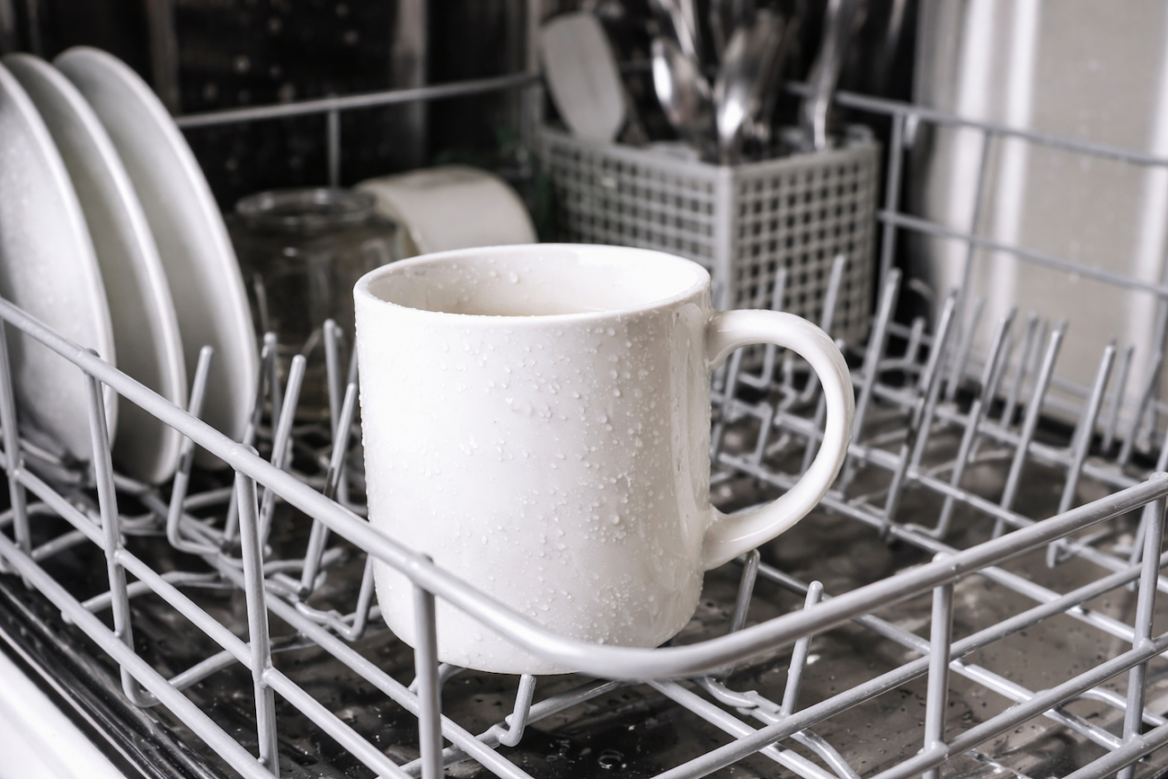 White ceramic mug placed in the lower rack of a dishwasher with water droplets on its surface