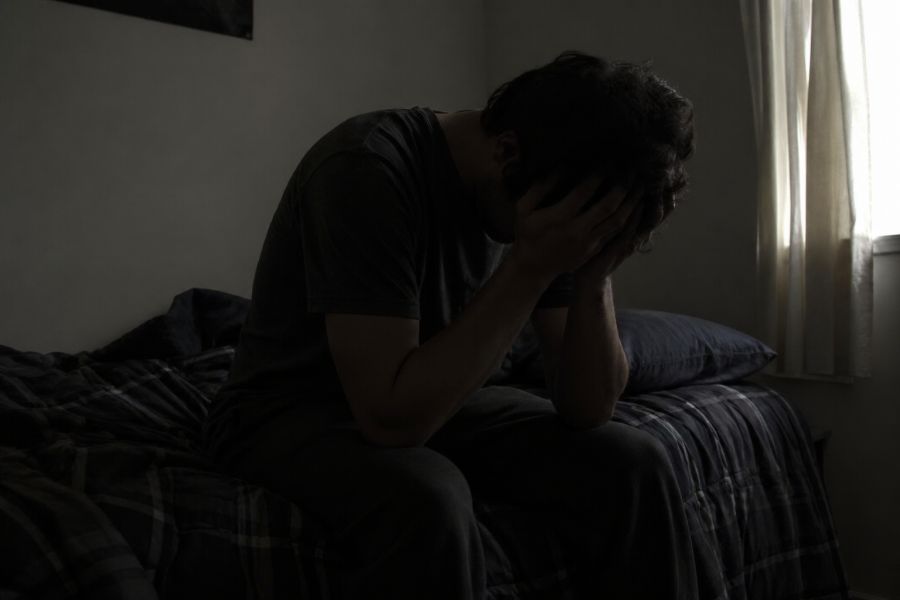 Depressed man sitting alone on a bed, holding his head in his hands in a dark room