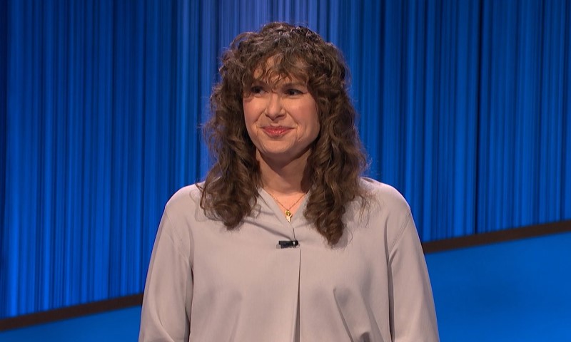 A woman with curly hair stands on a brightly lit game show set with a calm expression