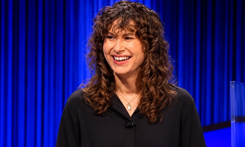 A person with curly brown hair smiles while wearing a black shirt on a bright blue-lit stage