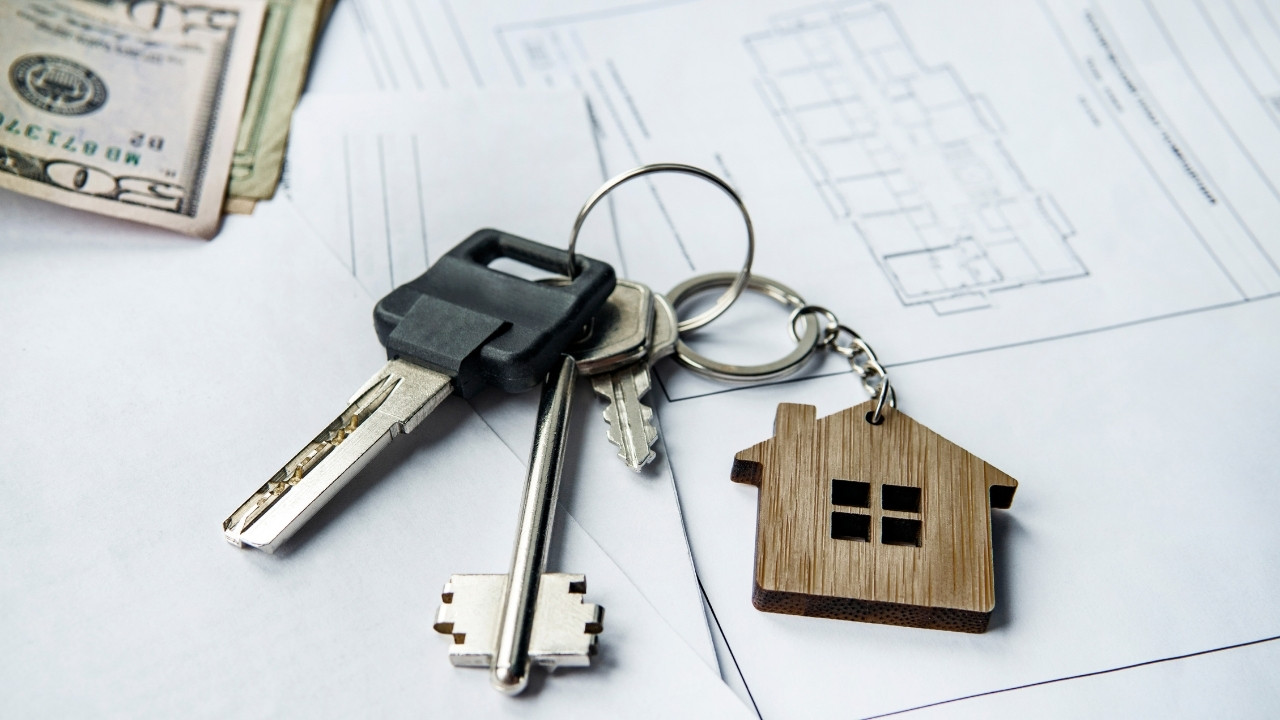 House keys and wooden house keychain resting on residential building plans beside cash.