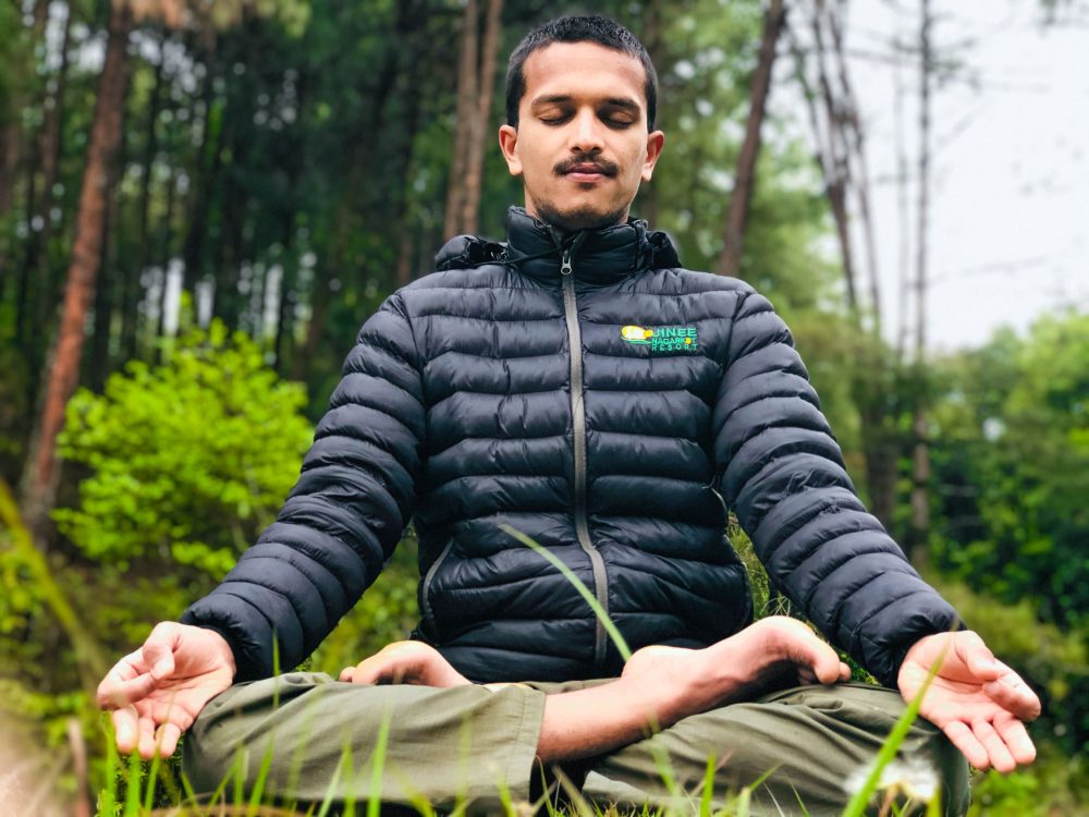 Barefoot man sitting in silent meditation pose with eyes closed near green woods