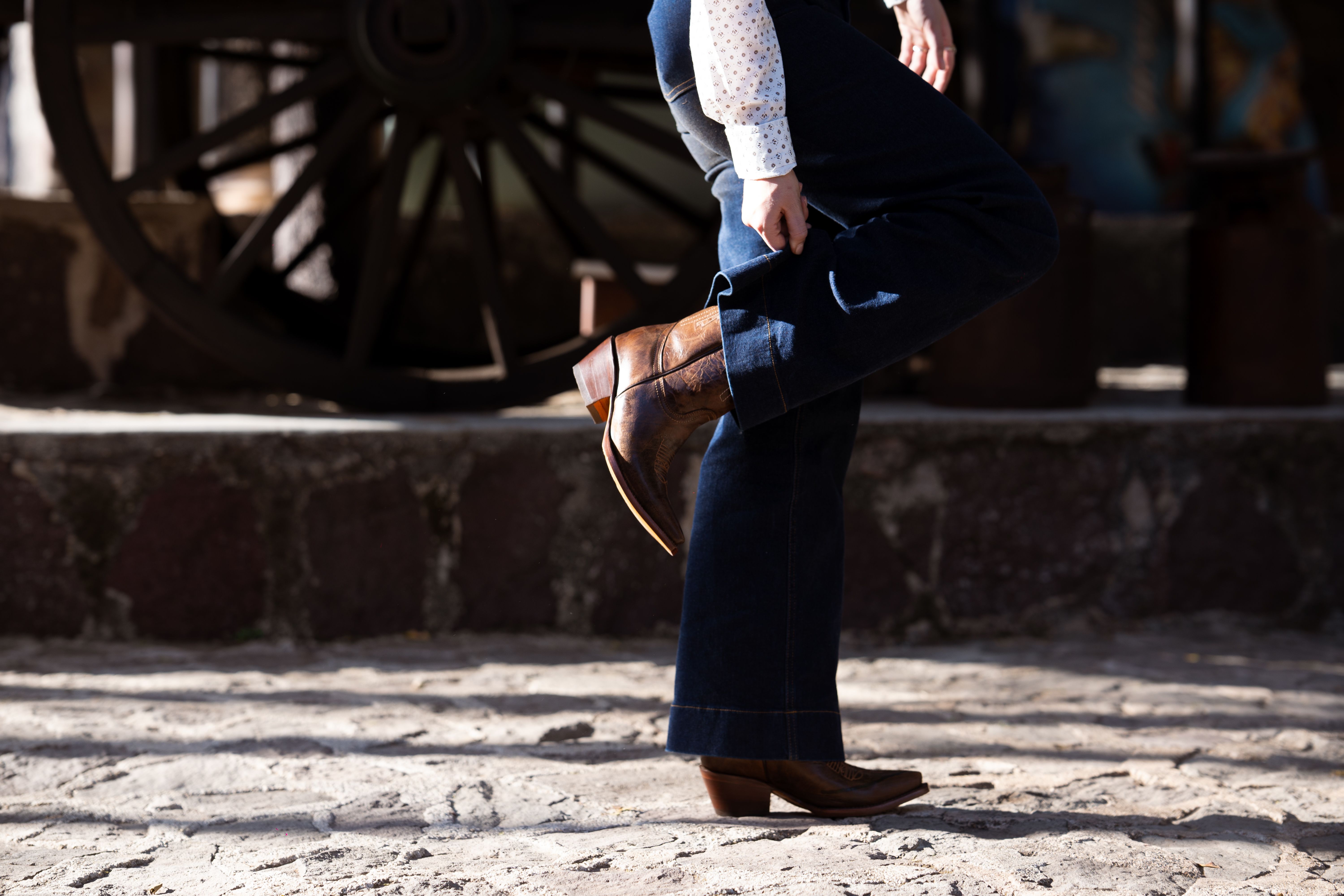 Person showcasing cowboy boots and jeans against a rustic vintage wagon backdrop