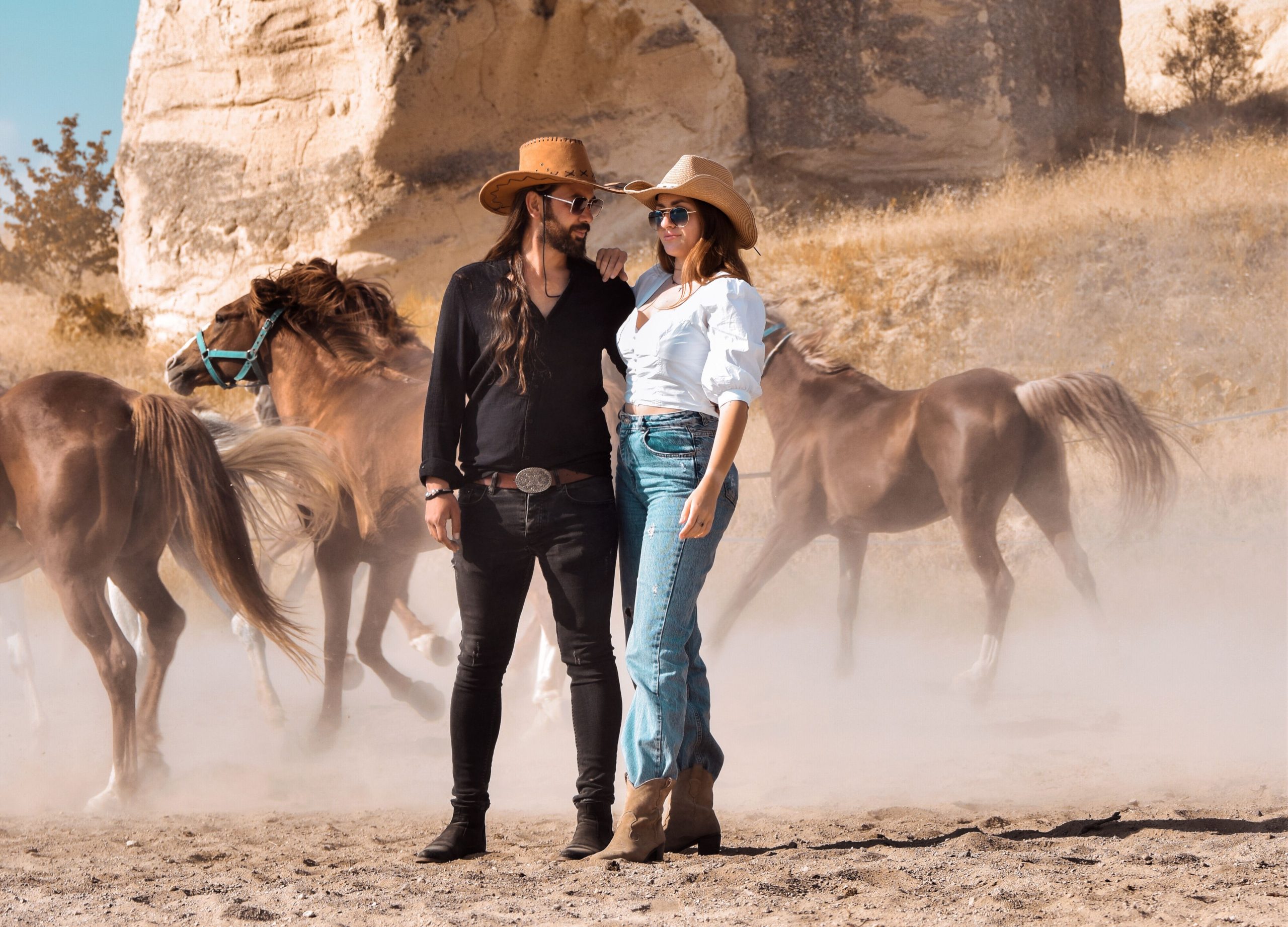 Fashionable couple in cowboy hats standing confidently with horses in a rustic outdoor setting