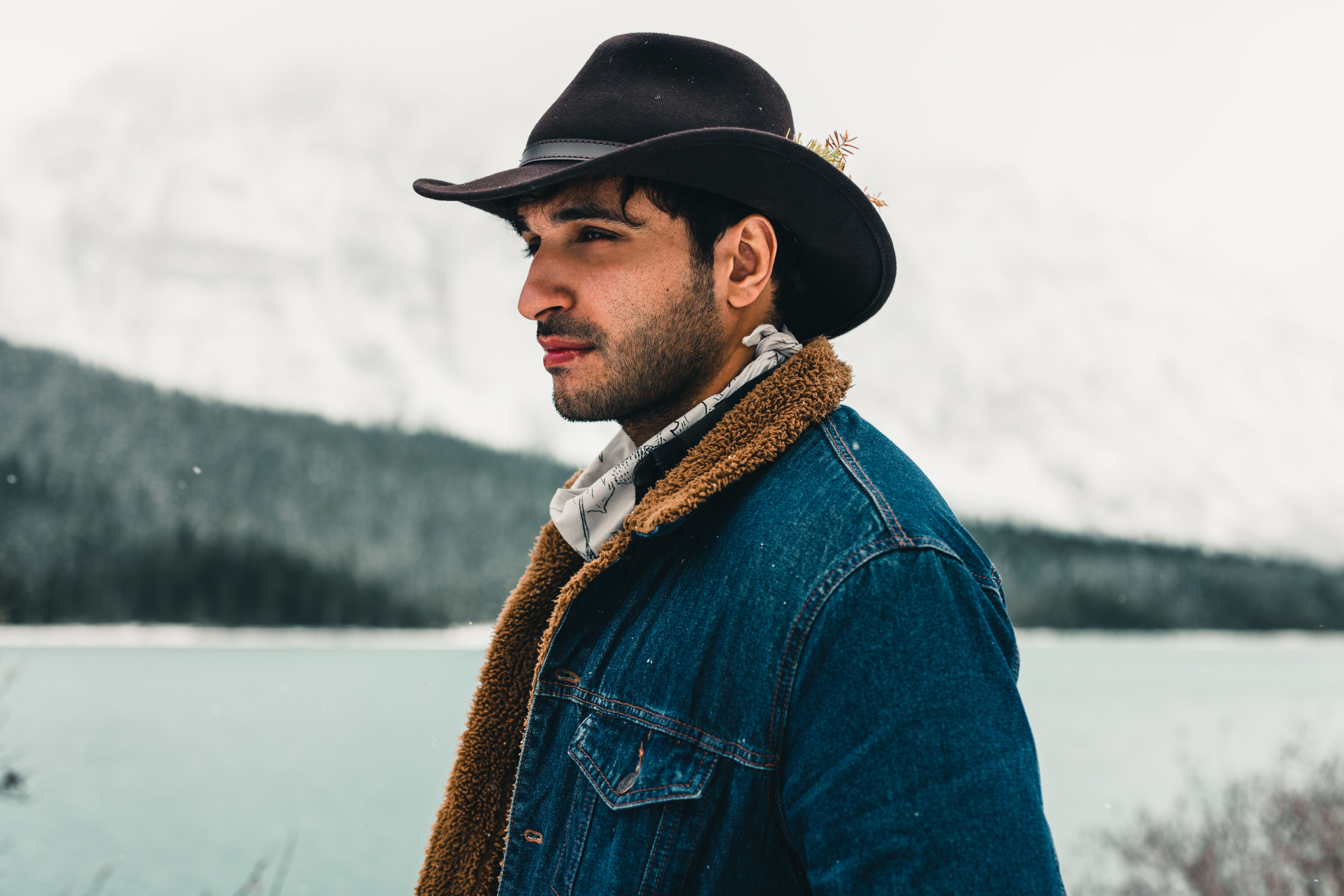 Man wearing a cowboy hat and denim jacket by a scenic lake