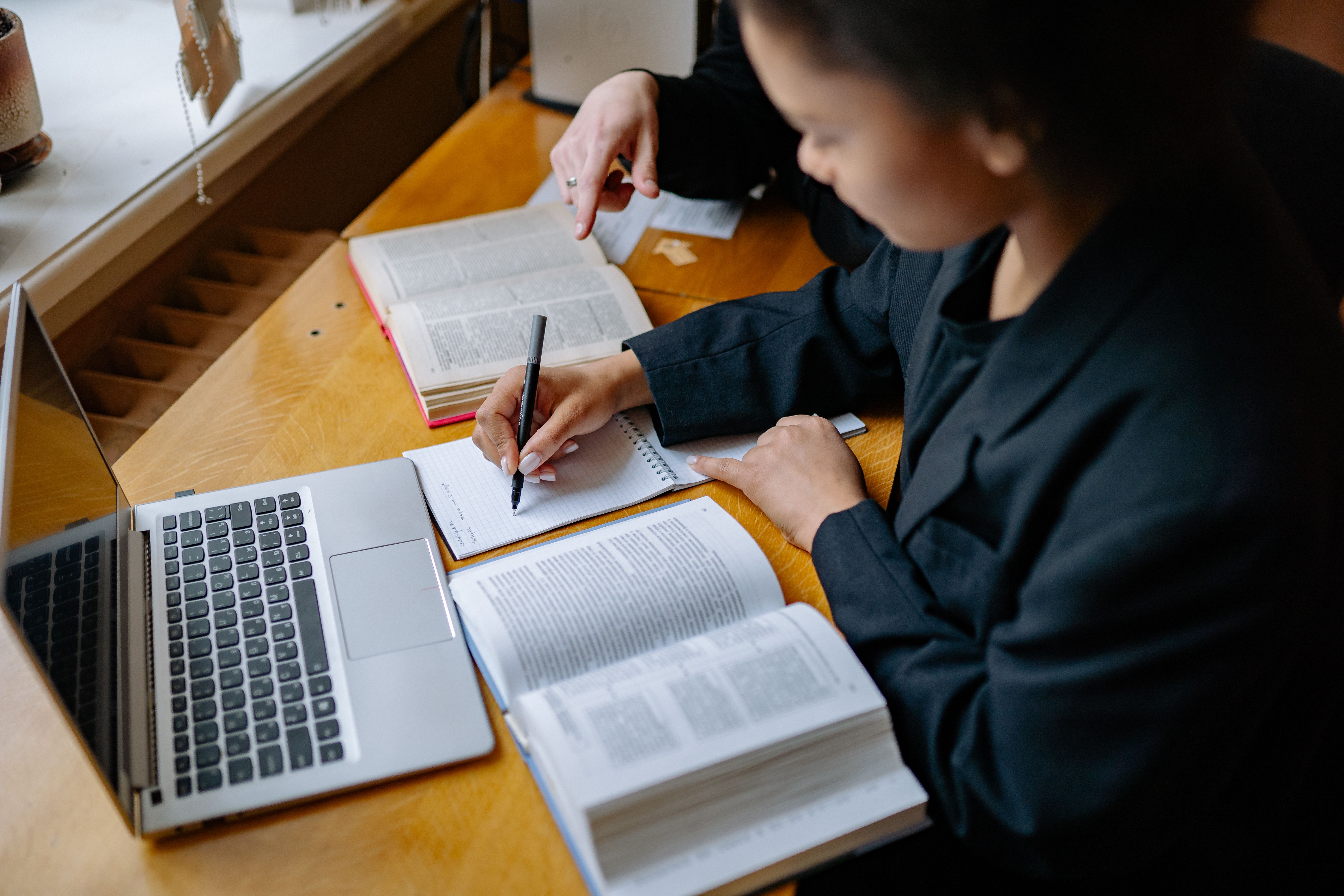 Two students taking notes with books and a laptop during a study session