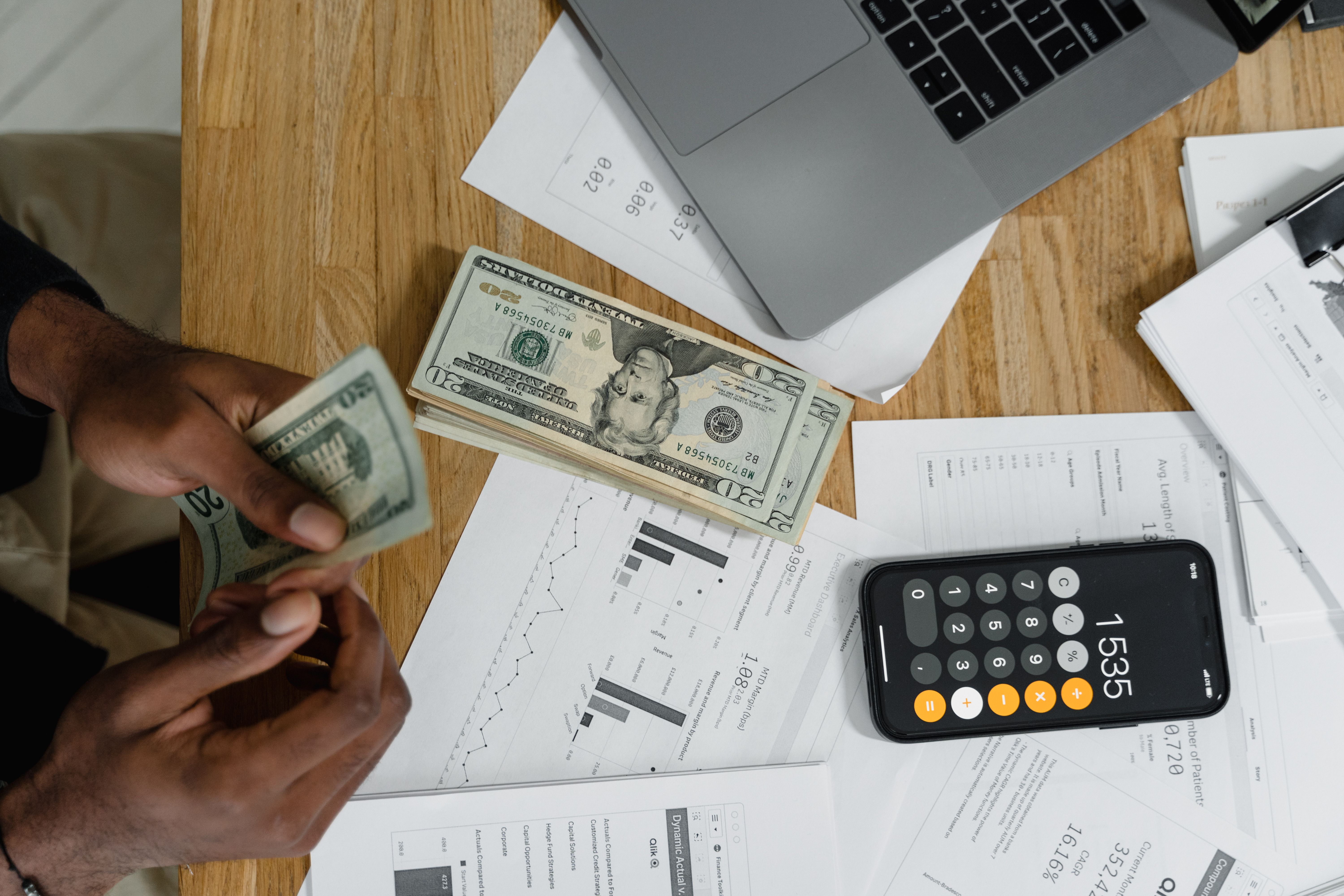Close-up of hands counting cash on desk with calculator, charts, and laptop